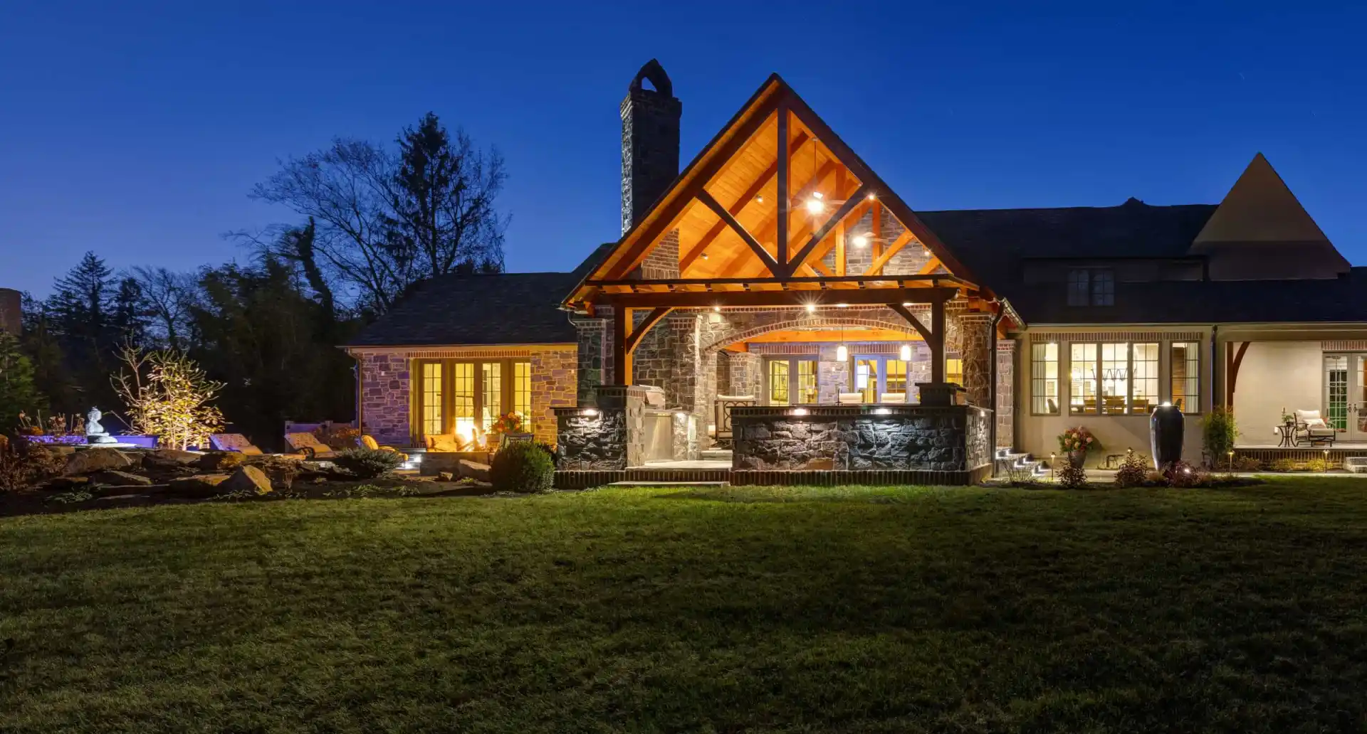A large stone house with a wooden patio is warmly lit against a twilight sky in Delaware County, PA. The patio features a fireplace and seating area, perfect for outdoor living. The surrounding yard boasts expert landscaping with trees and shrubs, enhancing its natural beauty.