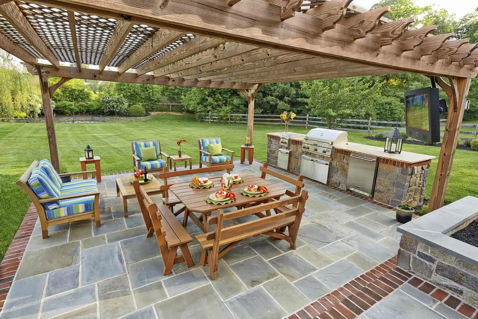 A spacious outdoor patio in Delaware County, PA, featuring a wooden pergola and stone flooring. Cushioned chairs and a bench circle a wooden table set with plates. A grill and countertop sit in the corner, surrounded by lush trees—a perfect example of expert outdoor living design.