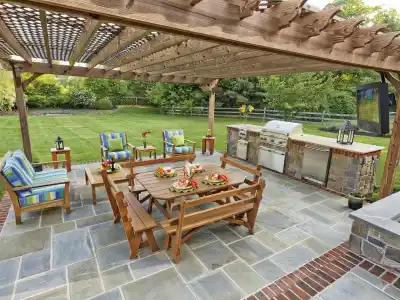 A spacious outdoor patio in Delaware County, PA, featuring a wooden pergola and stone flooring. Cushioned chairs and a bench circle a wooden table set with plates. A grill and countertop sit in the corner, surrounded by lush trees—a perfect example of expert outdoor living design.