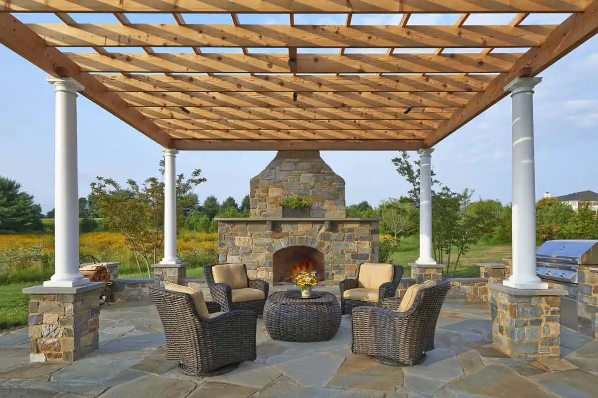 A stone patio with wicker chairs and a round table surrounds a stone fireplace under a wooden pergola supported by white columns, set in a landscaped garden.