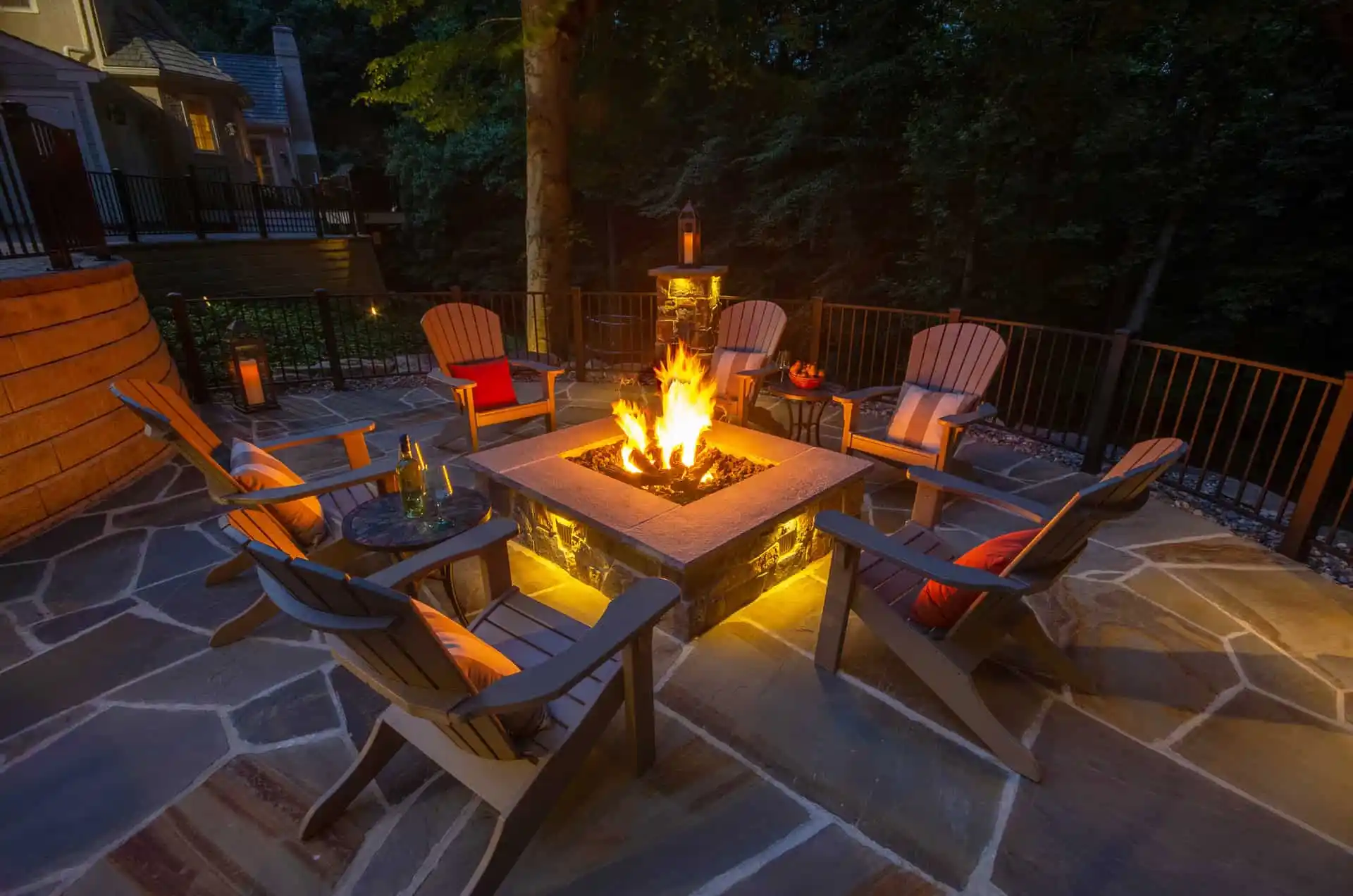 A cozy outdoor patio features a stone fire pit surrounded by six cushioned Adirondack chairs. Subtle lighting and the fire's glow illuminate the scene. With trees and a house in view, this serene evening ambiance highlights premium outdoor living in Delaware County, PA.