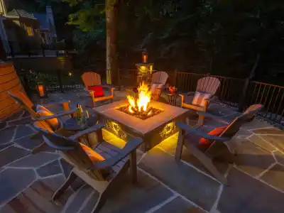 A cozy outdoor patio features a stone fire pit surrounded by six cushioned Adirondack chairs. Subtle lighting and the fire's glow illuminate the scene. With trees and a house in view, this serene evening ambiance highlights premium outdoor living in Delaware County, PA.