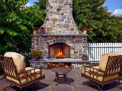 Outdoor living in Delaware County, PA, shines on this patio with a large stone fireplace at its center. A crackling fire warms two cushioned metal chairs and a small round table. Surrounded by lush trees and a white picket fence, it's the perfect blend of nature and design under a blue sky.