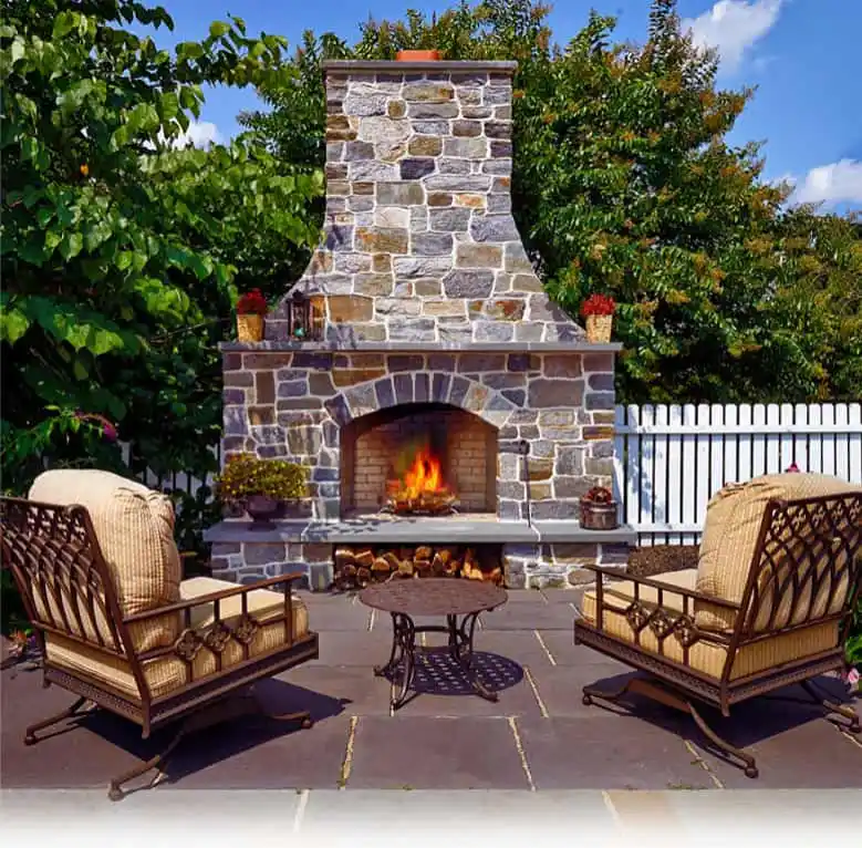 Outdoor living in Delaware County, PA, shines on this patio with a large stone fireplace at its center. A crackling fire warms two cushioned metal chairs and a small round table. Surrounded by lush trees and a white picket fence, it's the perfect blend of nature and design under a blue sky.