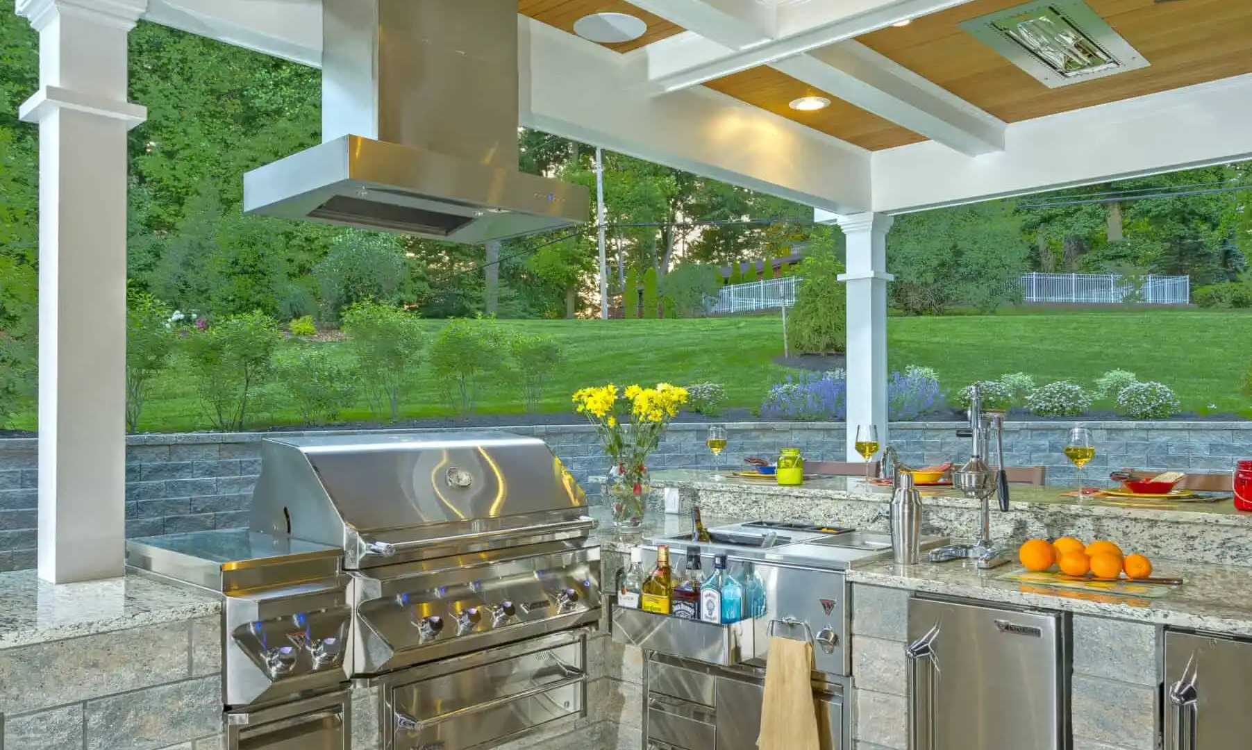 Experience the ultimate in outdoor living in Delaware County, PA, with this kitchen featuring a stainless steel grill, sink, and refrigerator under a wooden ceiling. A stone countertop displays vases with yellow flowers and fresh oranges. Greenery and a stone wall complete this serene setting.