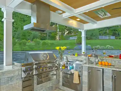 Experience the ultimate in outdoor living in Delaware County, PA, with this kitchen featuring a stainless steel grill, sink, and refrigerator under a wooden ceiling. A stone countertop displays vases with yellow flowers and fresh oranges. Greenery and a stone wall complete this serene setting.