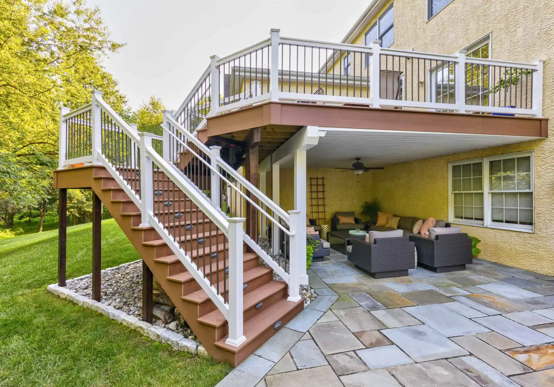 Outdoor view of a two-story deck featuring a staircase with white railings, leading to a ground-level patio. With wicker furniture and lush landscaping services Delaware County PA, this outdoor oasis is perfect for enjoying nature's beauty amidst the greenery and manicured lawn.