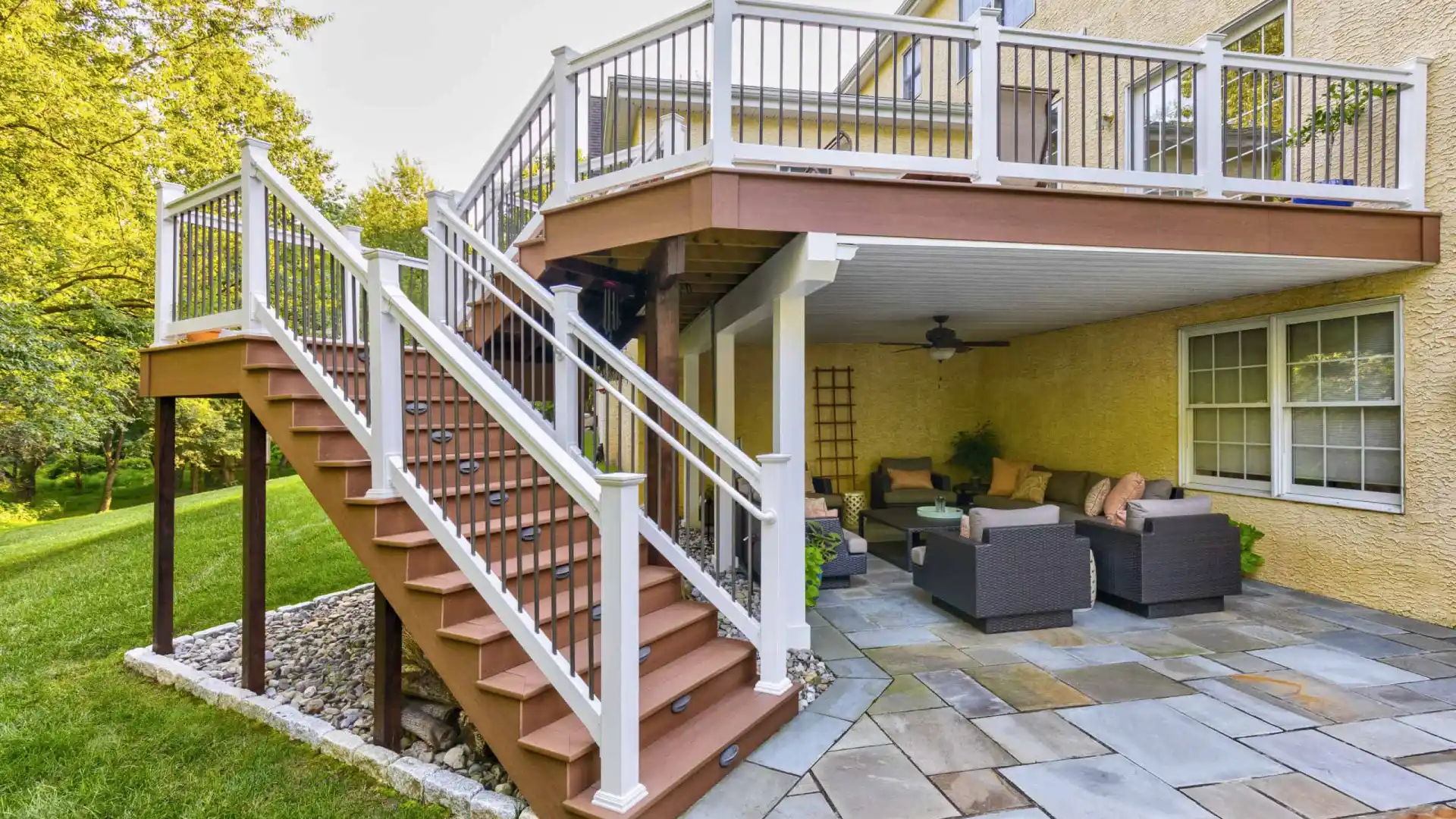 Outdoor view of a two-story deck featuring a staircase with white railings, leading to a ground-level patio. With wicker furniture and lush landscaping services Delaware County PA, this outdoor oasis is perfect for enjoying nature's beauty amidst the greenery and manicured lawn.