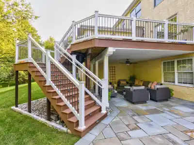 Outdoor view of a two-story deck featuring a staircase with white railings, leading to a ground-level patio. With wicker furniture and lush landscaping services Delaware County PA, this outdoor oasis is perfect for enjoying nature's beauty amidst the greenery and manicured lawn.