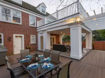 A spacious outdoor patio features a dining table set with blue and white dishware, chairs, a built-in grill, and a covered lounge area. Expertly crafted for outdoor living in Delaware County PA, the patio is attached to a brick house with large windows and a white balcony above.