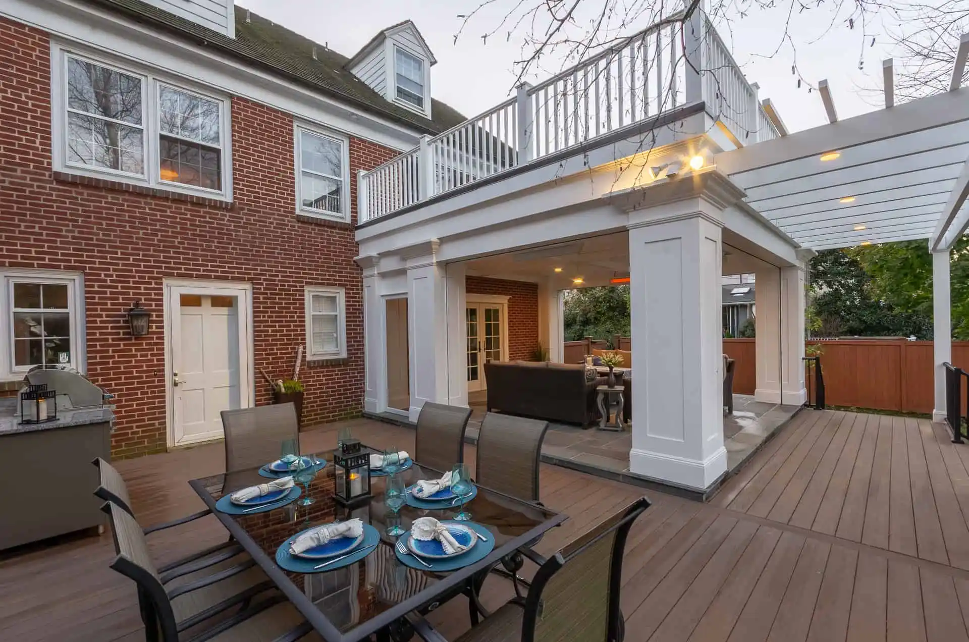 A spacious outdoor patio features a dining table set with blue and white dishware, chairs, a built-in grill, and a covered lounge area. Expertly crafted for outdoor living in Delaware County PA, the patio is attached to a brick house with large windows and a white balcony above.