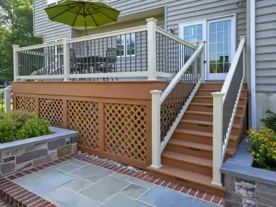 A wooden deck is attached to a house, featuring white railings and stairs leading down to a stone patio. Perfect for outdoor living in Delaware County, PA, it boasts a dining table with chairs and a green umbrella. Flowering plants add charm beside the stairs.