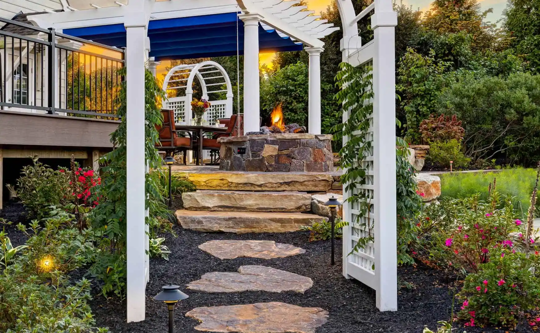 A garden at sunset in Delaware County, PA, features a stone pathway leading through a white arbor draped with climbing plants. The path ascends to a fire pit surrounded by chairs, nestled among lush greenery. The colorful sky and expertly designed landscaping create the perfect outdoor living space.
