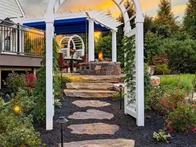 A garden at sunset in Delaware County, PA, features a stone pathway leading through a white arbor draped with climbing plants. The path ascends to a fire pit surrounded by chairs, nestled among lush greenery. The colorful sky and expertly designed landscaping create the perfect outdoor living space.