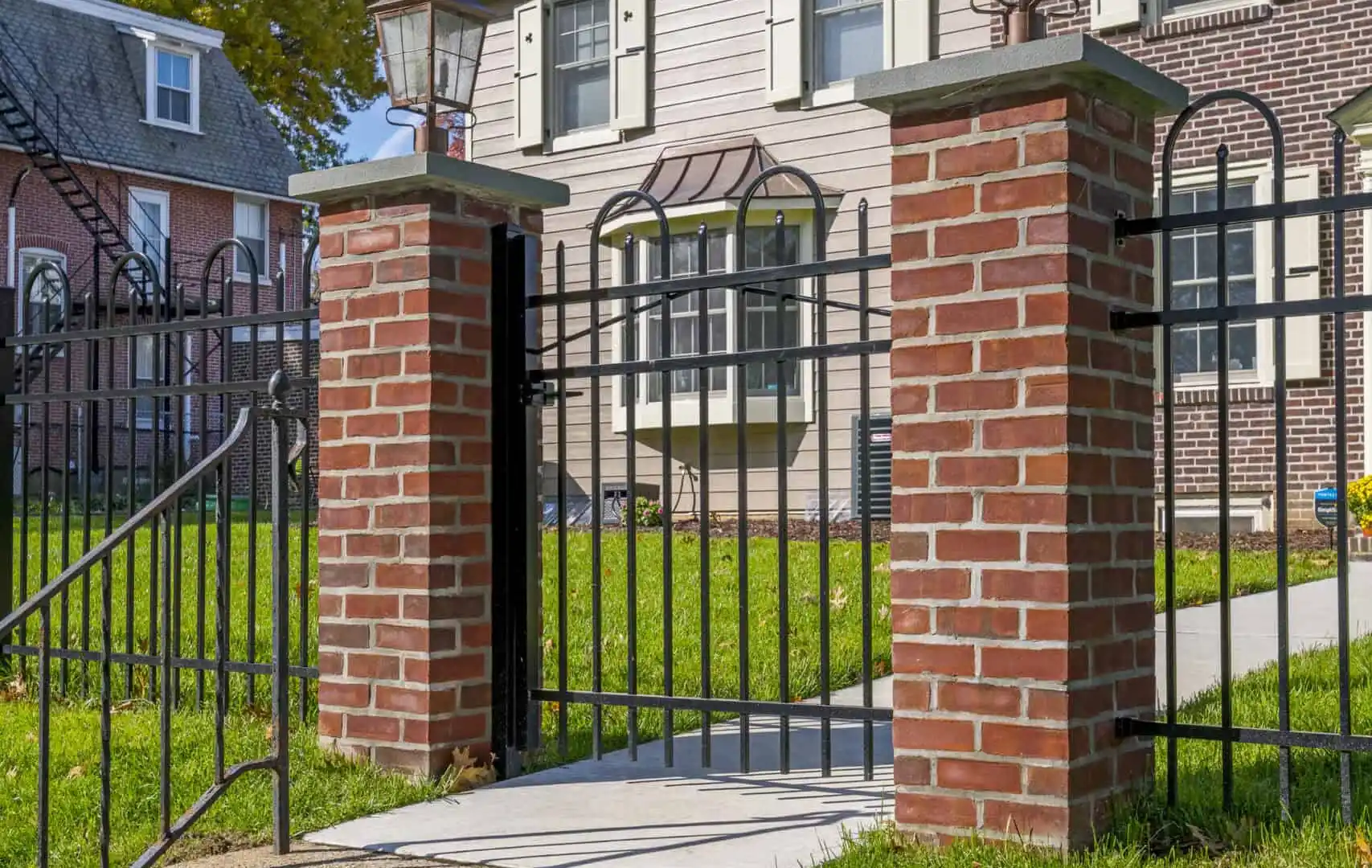 A bright, sunny day showcases a two-story house with a brick facade and white siding. In the foreground, expertly crafted hardscaping services in Delaware County, PA bring charm to the brick and wrought iron gate leading up stone steps, surrounded by green grass and a nearby house partially visible.