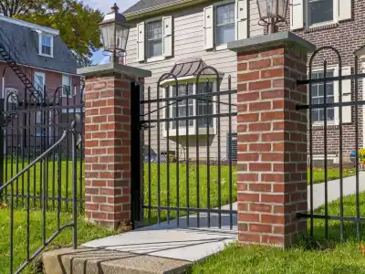 A bright, sunny day showcases a two-story house with a brick facade and white siding. In the foreground, expertly crafted hardscaping services in Delaware County, PA bring charm to the brick and wrought iron gate leading up stone steps, surrounded by green grass and a nearby house partially visible.