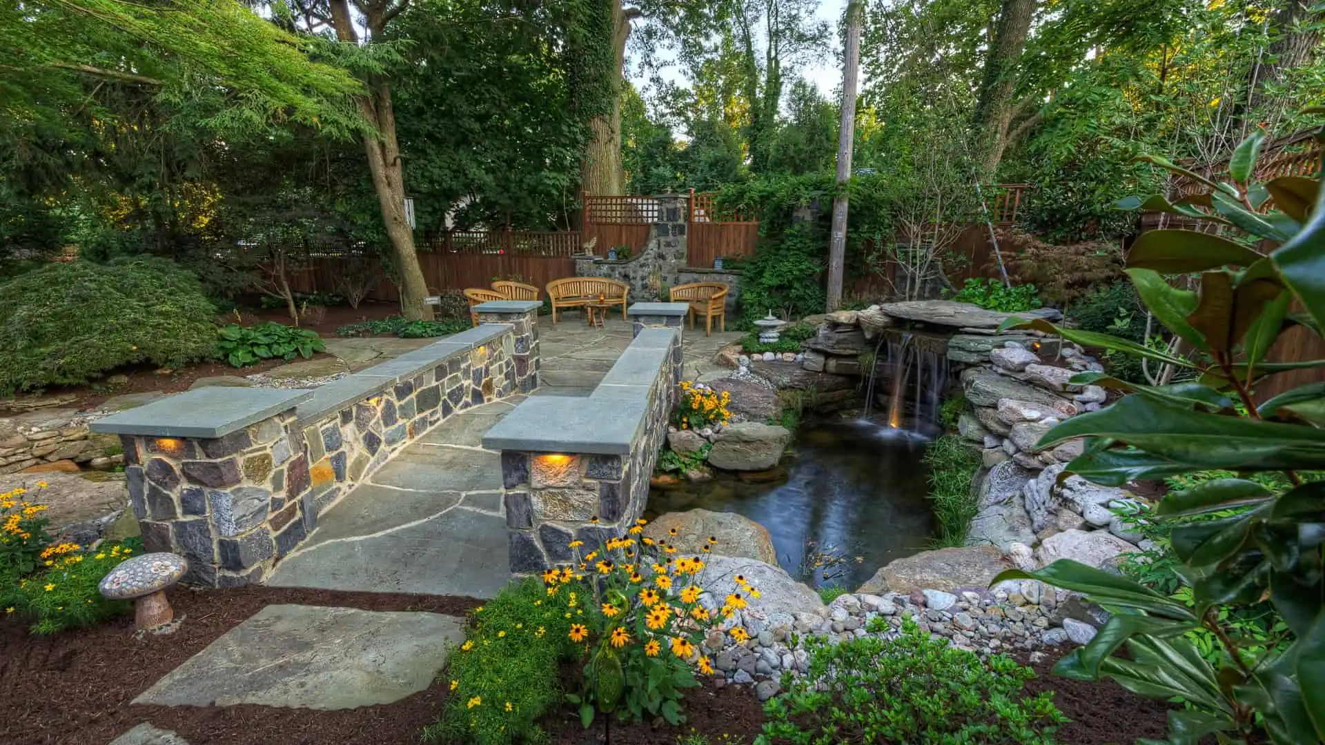 A serene garden with a stone path leading to a small waterfall and pond, this tranquil outdoor living space in Delaware County, PA, is surrounded by lush greenery and yellow flowers. Wooden chairs and a fence complete the scene under a canopy of trees.