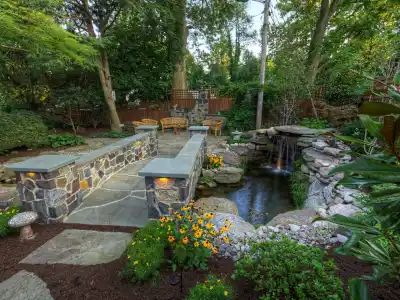 A serene garden with a stone path leading to a small waterfall and pond, this tranquil outdoor living space in Delaware County, PA, is surrounded by lush greenery and yellow flowers. Wooden chairs and a fence complete the scene under a canopy of trees.