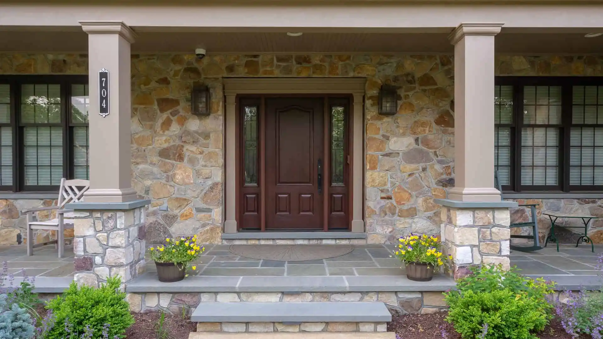 A stone-front porch in Delaware County, PA, boasts a dark wood door flanked by two windows. Two potted plants with yellow flowers enhance its charm. A chair and small table offer outdoor living comfort on the left. Slate steps lead up as house number 