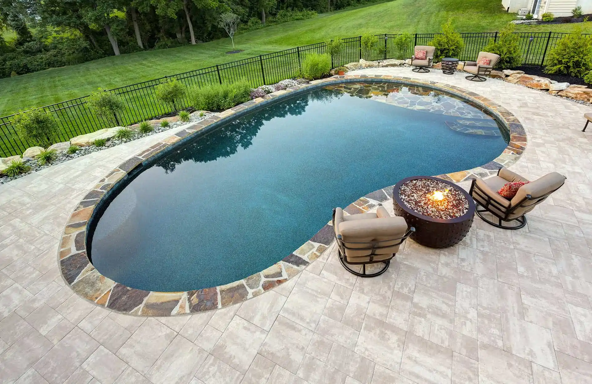 An oval swimming pool with stone edging sits in a backyard with a tile patio, surrounded by green grass and trees. Two cushioned chairs face a round fire pit on the patio near the pool.