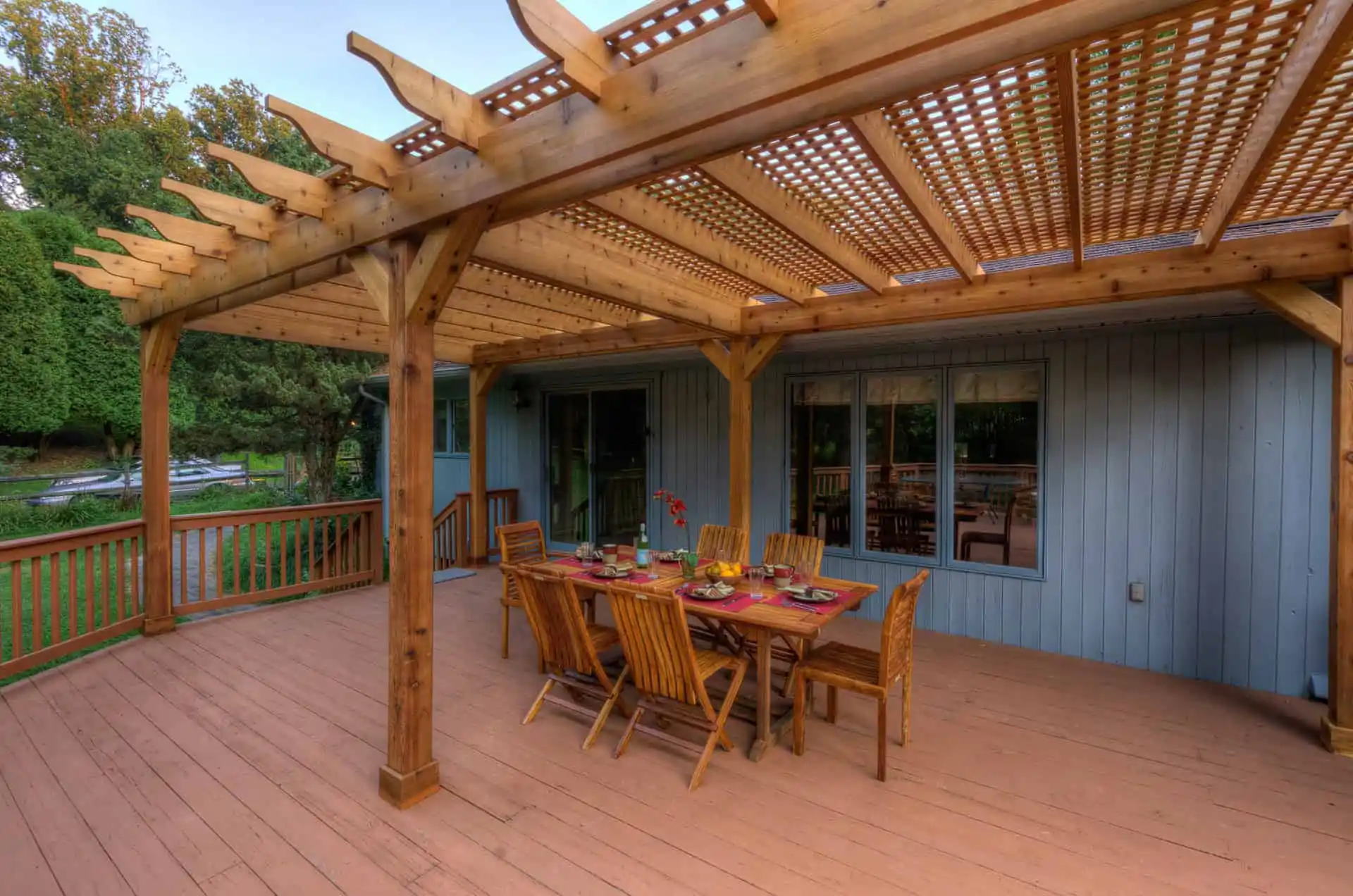 A wooden deck with a pergola overhead features a dining table set for eight, surrounded by wooden chairs. The deck overlooks a green yard and is attached to a blue house with large windows.