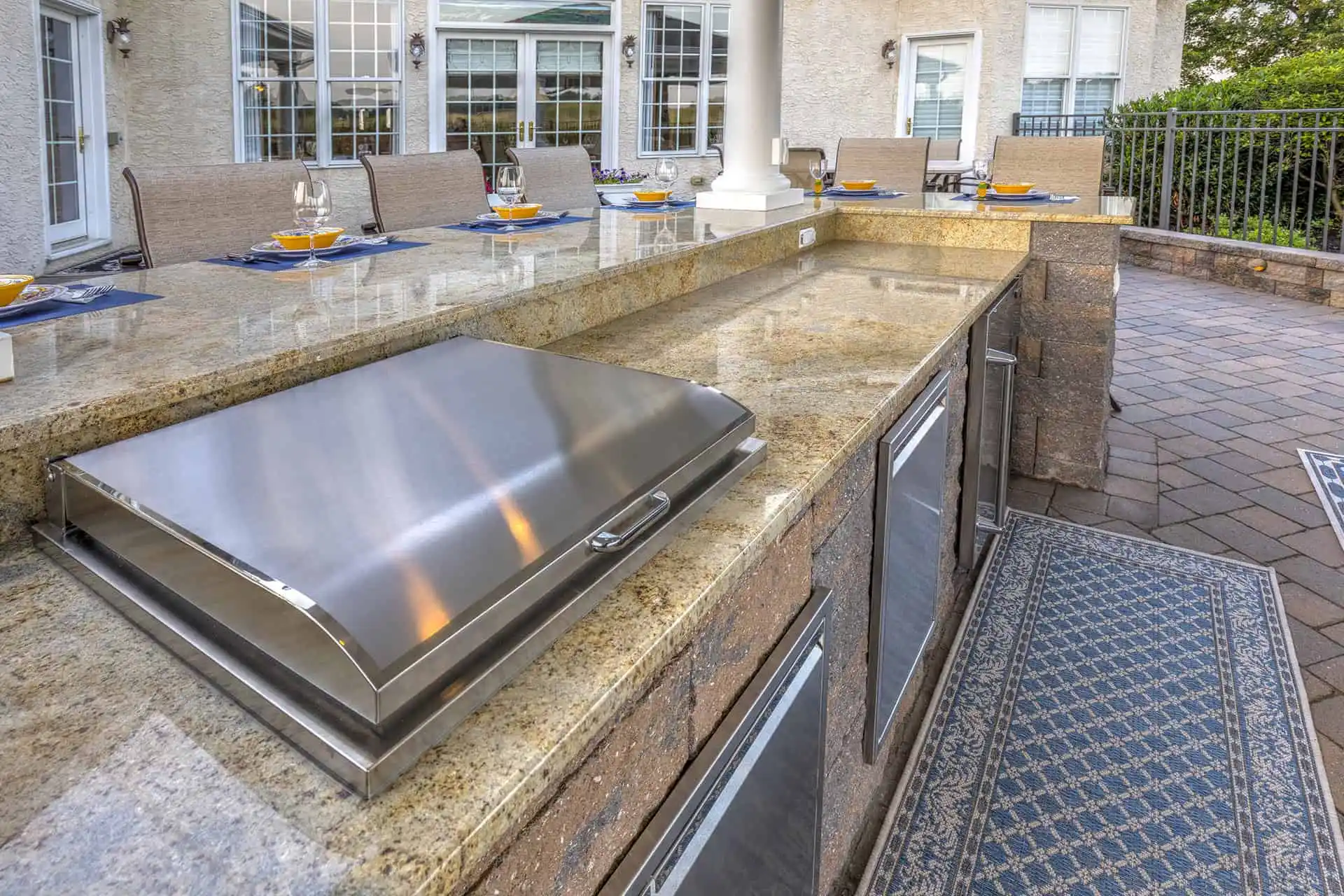 Outdoor kitchen with a stone countertop, built-in grill, stainless steel appliances, and a dining area set with plates and glasses. The space overlooks a patio with pavers and outdoor seating.