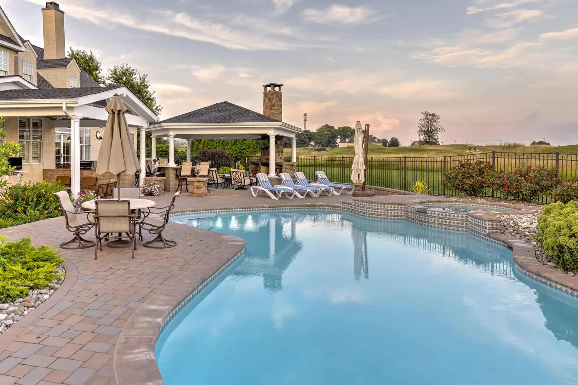 A backyard with a large swimming pool, surrounded by lounge chairs, patio tables with umbrellas, landscaped plants, and a covered outdoor seating area near a fireplace. The sky is partly cloudy at sunset.