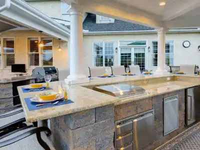 A modern outdoor kitchen and dining area with granite countertops, a built-in grill, stainless steel appliances, and a table set for six with yellow plates and blue placemats under a covered patio—perfect for fans of stylish outdoor kitchens.