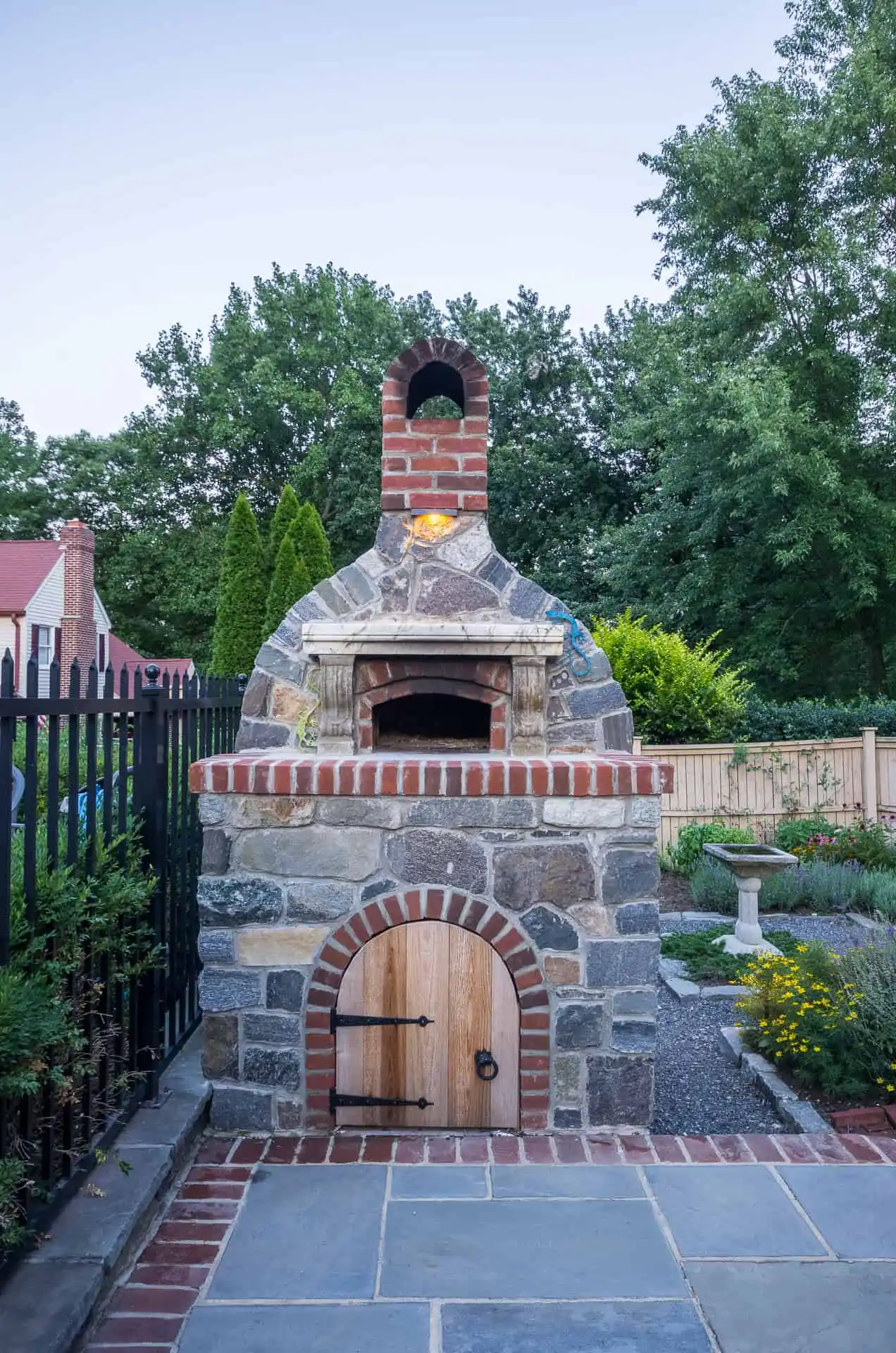 A stone and brick outdoor pizza oven with a wooden door sits on a patio, surrounded by a fence, garden, and trees in a backyard setting.