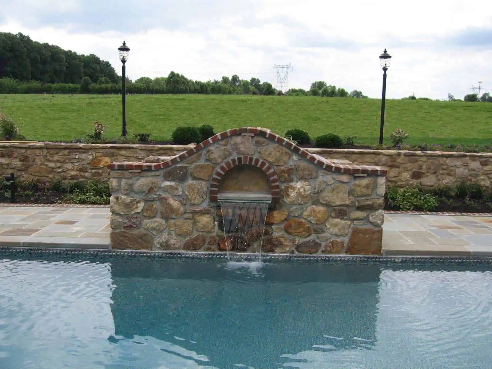 A stone wall with a small arched fountain spilling water into a swimming pool, set outdoors with grassy fields, trees, and two lamp posts in the background.