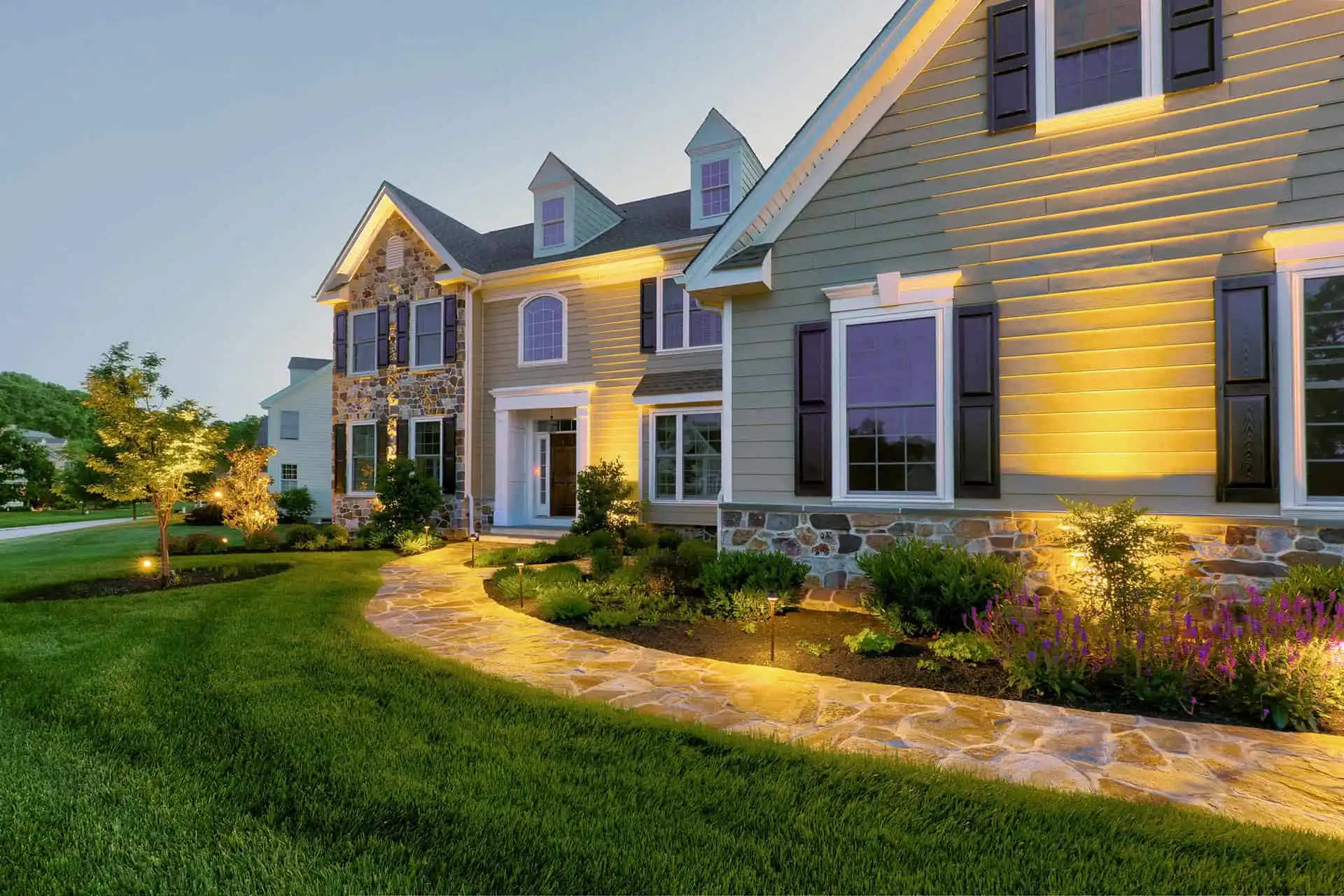 A large suburban house with stone and siding exterior, lit by warm outdoor lighting. A stone walkway curves through a well-manicured lawn and landscaped bushes, with the sky transitioning to dusk.