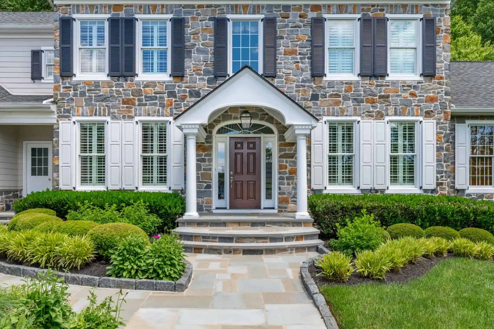 Stone house facade with white window shutters and a dark wooden door, surrounded by well-maintained greenery and a small garden. Expert landscaping services in Delaware County, PA ensure the entrance's semi-circular stone staircase and triangular pediment shine elegantly above the door.