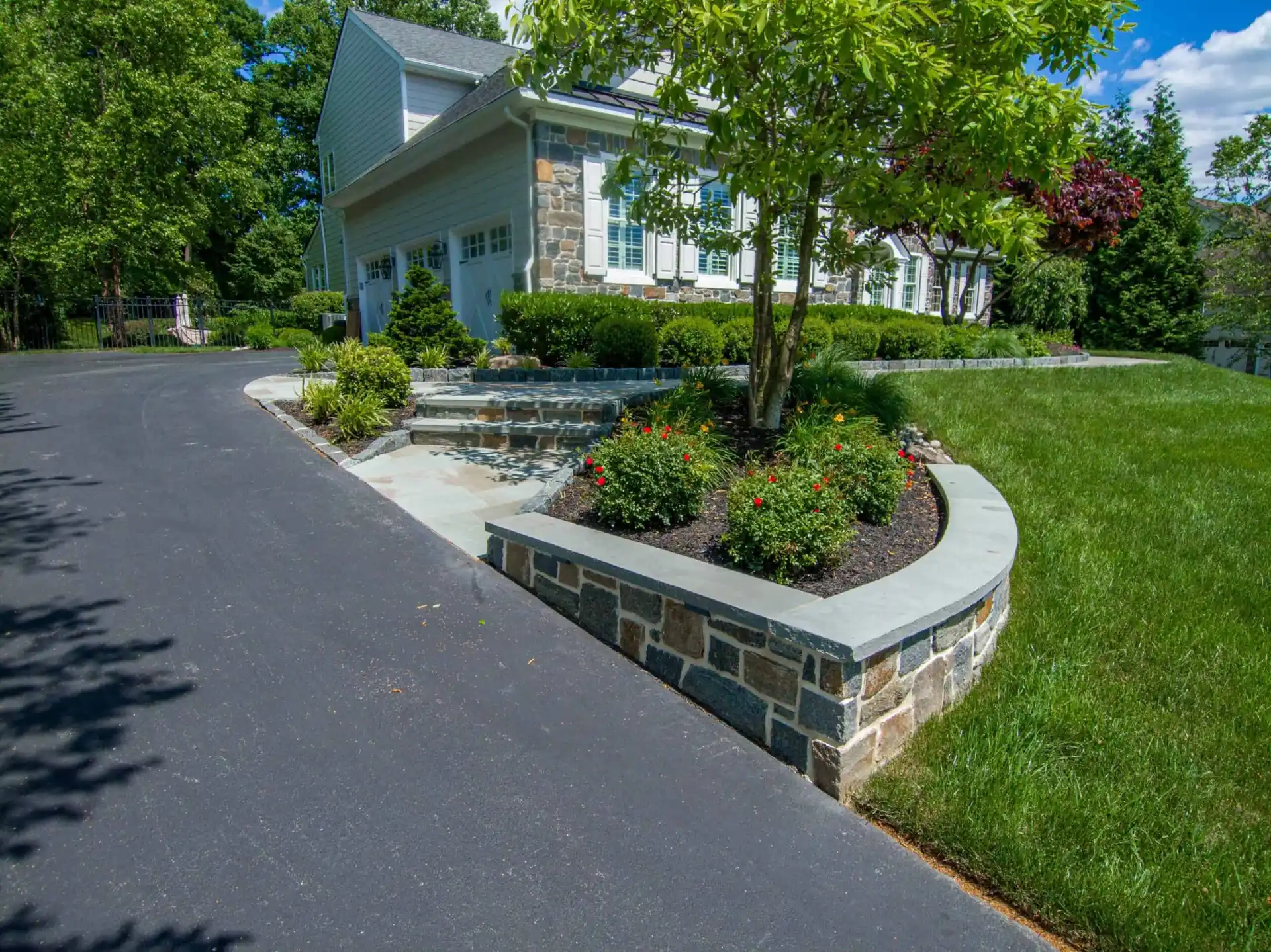 A well-maintained suburban home with gray stone exterior and white trim, nestled in Delaware County PA. The house is surrounded by lush greenery, a stone wall, manicured bushes, and colorful flower beds—all enhanced by expert landscaping services. A curved black asphalt driveway leads up to the garage.