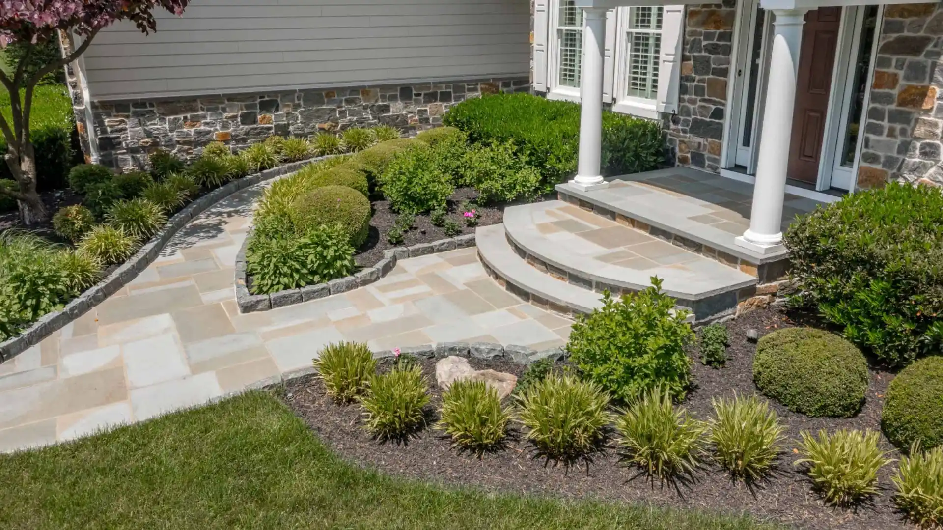 A well-maintained garden with a curved stone pathway leading to a house entrance showcases professional landscaping services in Delaware County, PA. Neatly trimmed bushes and small plants line the path and steps, while stone accents and white pillars frame the front door beautifully.