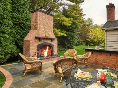 A cozy outdoor patio in Delaware County, PA, features a large brick fireplace with a roaring fire, surrounded by wicker chairs. A black metal table with plates and drinks sits nearby. Lush greenery enhances the inviting atmosphere, showcasing expert hardscaping services.