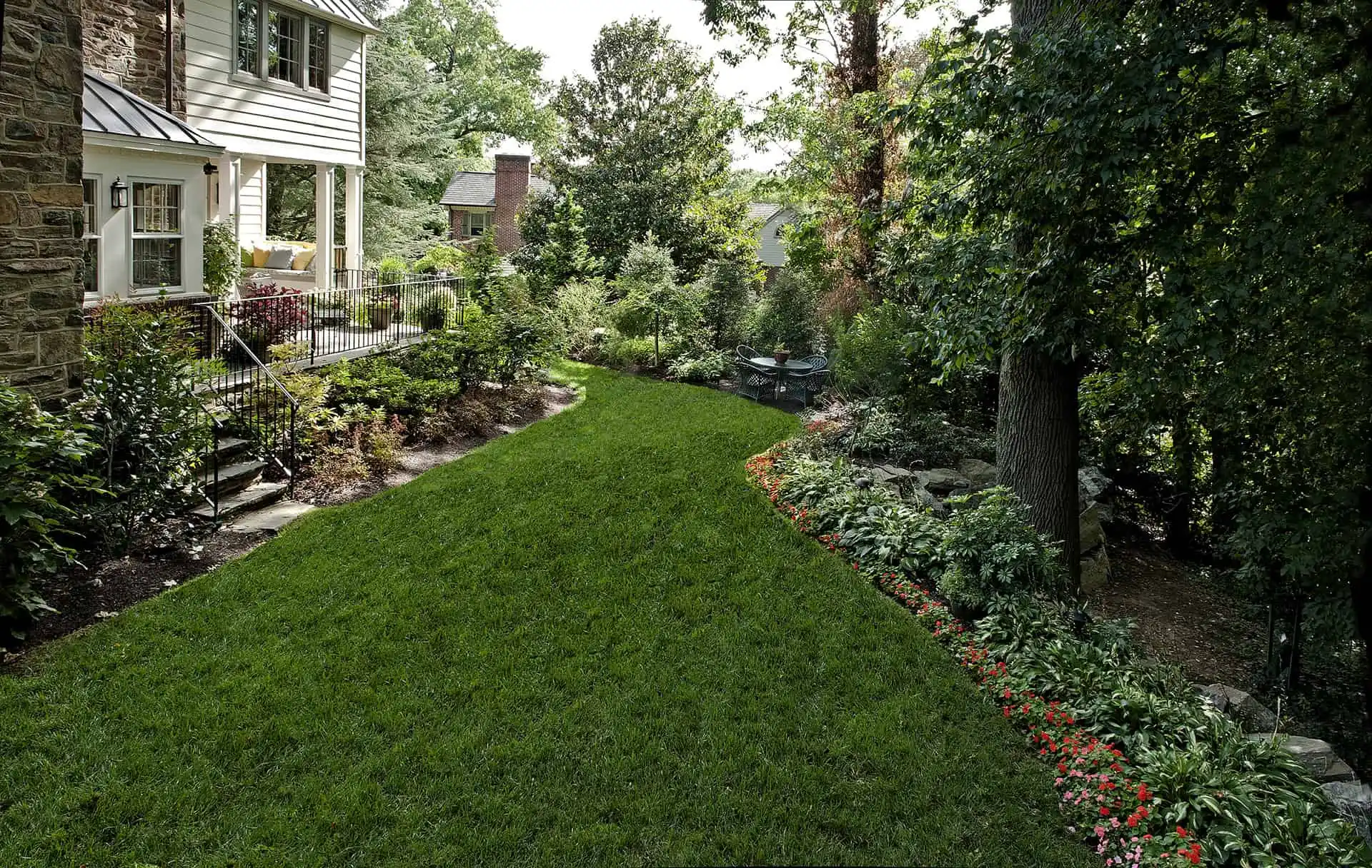 A lush green backyard features manicured grass, vibrant flowerbeds along a stone path, and a patio area adjoining a house with stone and siding, bordered by leafy trees and shrubs.