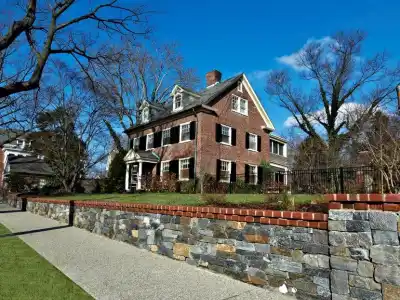 A large, three-story brick house with a steep roof and dormer windows stands behind a stone wall, perfectly complemented by professional landscaping services in Delaware County, PA. A sidewalk and grass lawn are in the foreground. Leafless trees frame the house under a clear blue sky.