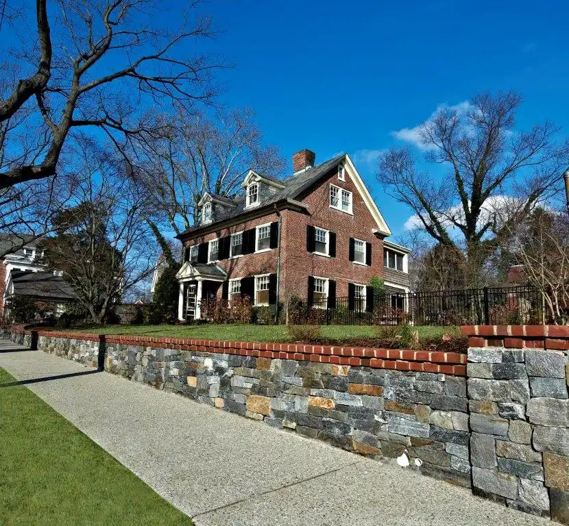 A large, three-story brick house with a steep roof and dormer windows stands behind a stone wall, perfectly complemented by professional landscaping services in Delaware County, PA. A sidewalk and grass lawn are in the foreground. Leafless trees frame the house under a clear blue sky.
