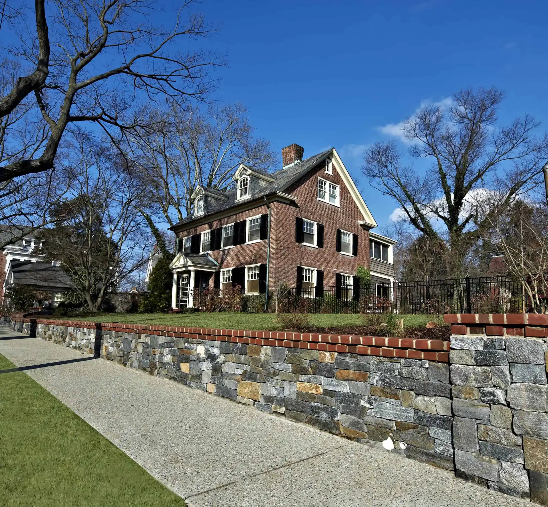 A large, three-story brick house with white trim sits behind a stone wall and iron fence on a suburban street, with leafless trees and a clear blue sky in the background.