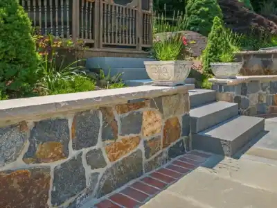 Image of a garden area with a stone retaining wall and stairs leading up to a wooden railing, showcasing expert hardscaping services in Delaware County, PA. Decorative stone planters with green foliage adorn the wall, while lush greenery and paved stones complete this outdoor living space.