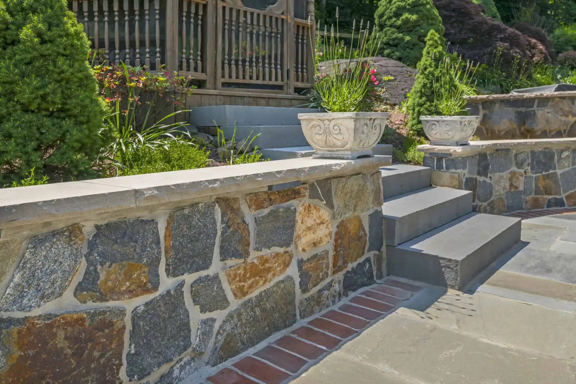 A stone garden wall and steps with gray pavers lead up to a wooden railing. Decorative stone planters with green plants sit on the wall, surrounded by lush greenery and landscaping.