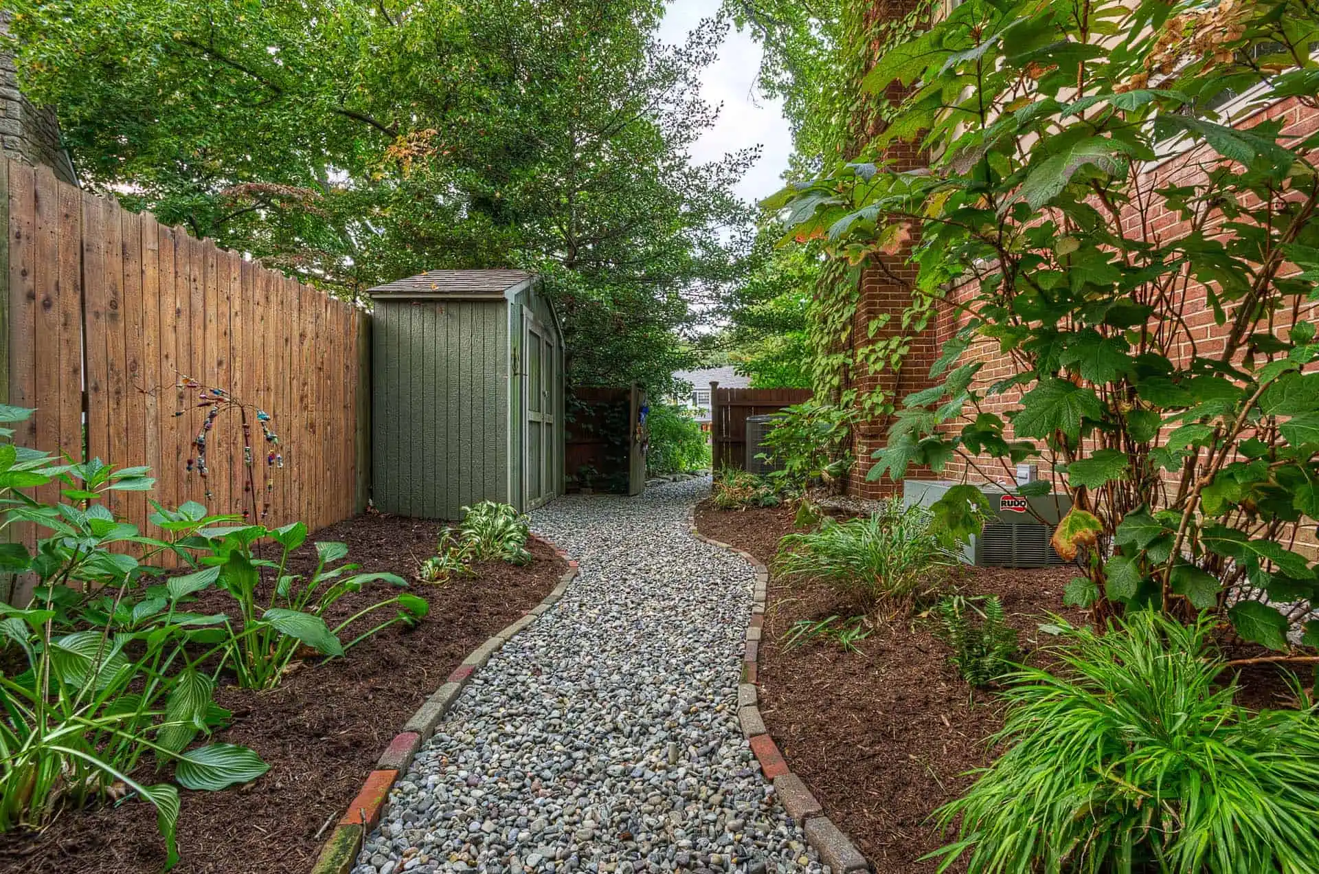 A curved pebble pathway runs between a wooden fence and a brick house, bordered by green plants and mulch, leading to a small green shed under leafy trees.