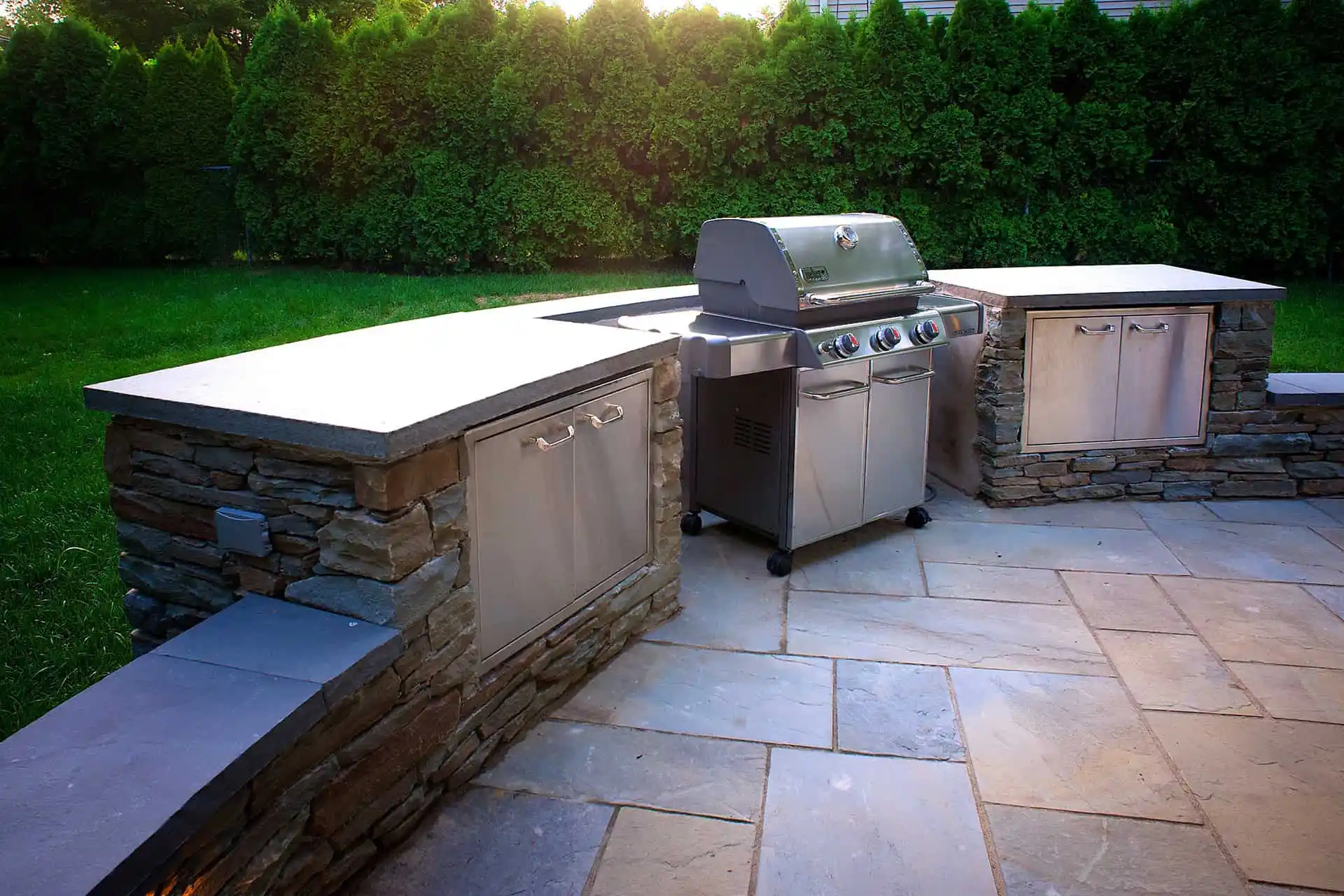 Outdoor kitchen with a built-in stainless steel grill, stone countertops, and storage cabinets, set on a stone patio with green grass and tall hedges in the background.