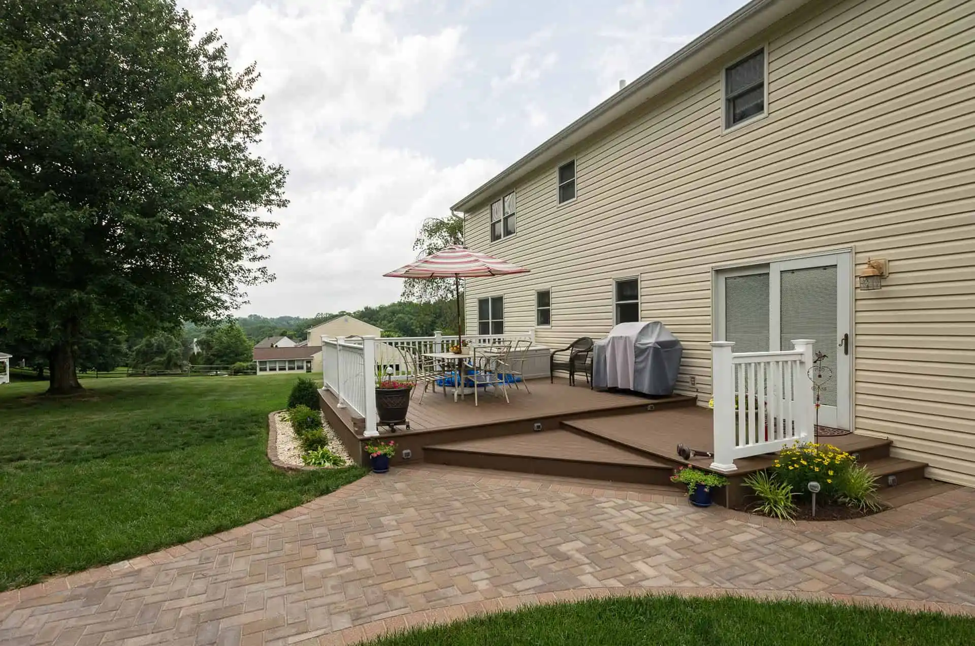 A backyard deck attached to a beige house features outdoor furniture, a grill, a striped umbrella, potted plants, and is surrounded by a lawn and a curved stone walkway.