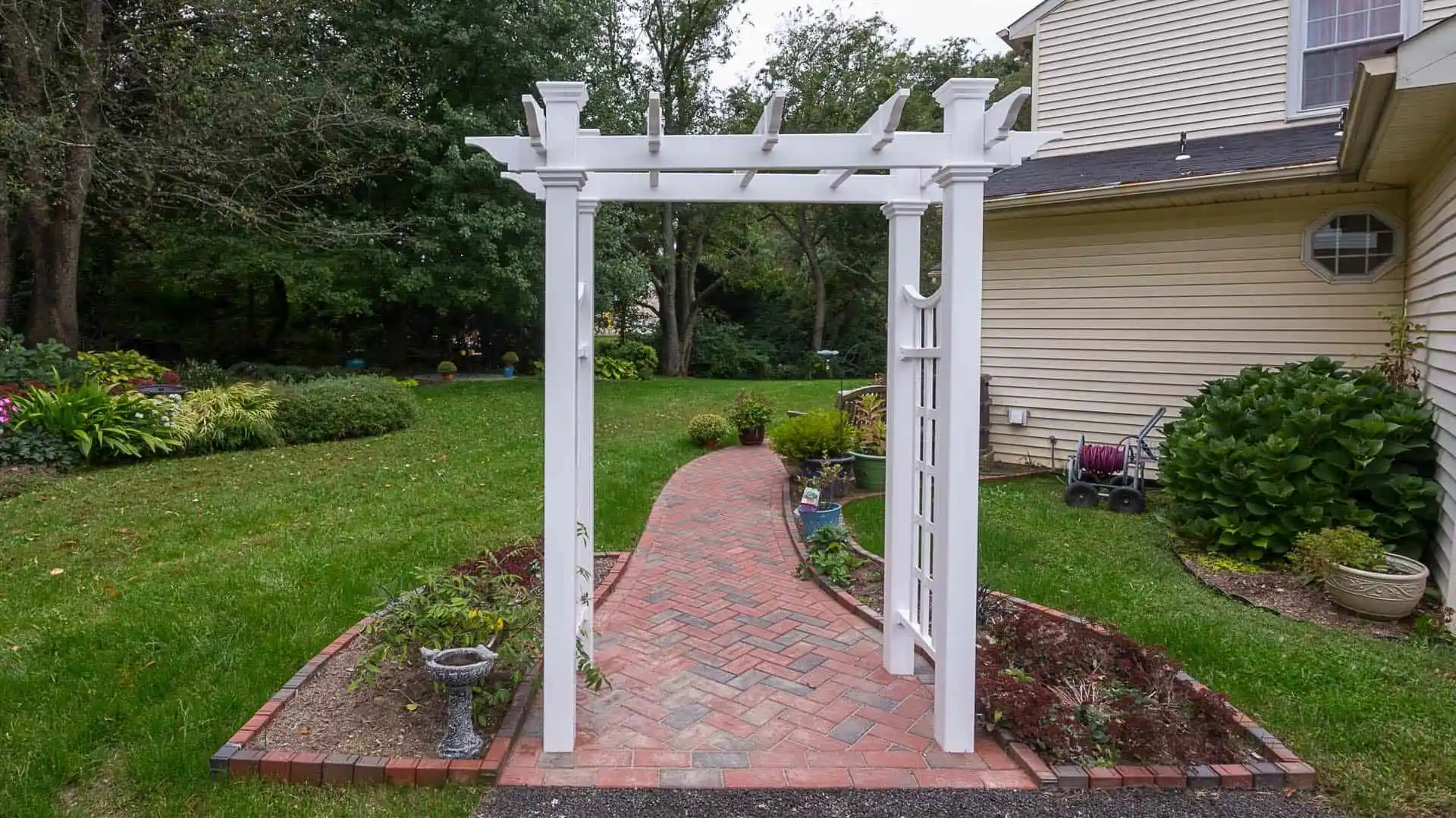 A white garden arbor with a lattice design stands at the entrance of a brick path, exemplifying expert landscaping services in Delaware County, PA. The path is bordered by small plants and leads to a lawn with trees in the background. A beige house is on the right side of the image