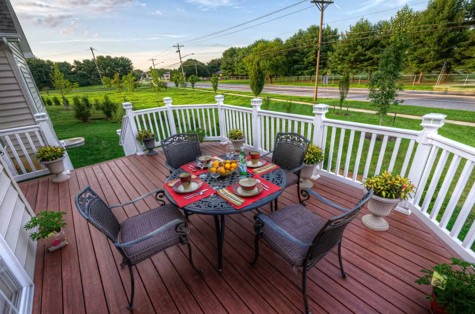 A wooden backyard deck with a black metal table and five chairs, set for a meal with red placemats, plates, cups, and a fruit bowl. Potted plants decorate the deck, with greenery and a road in the background.