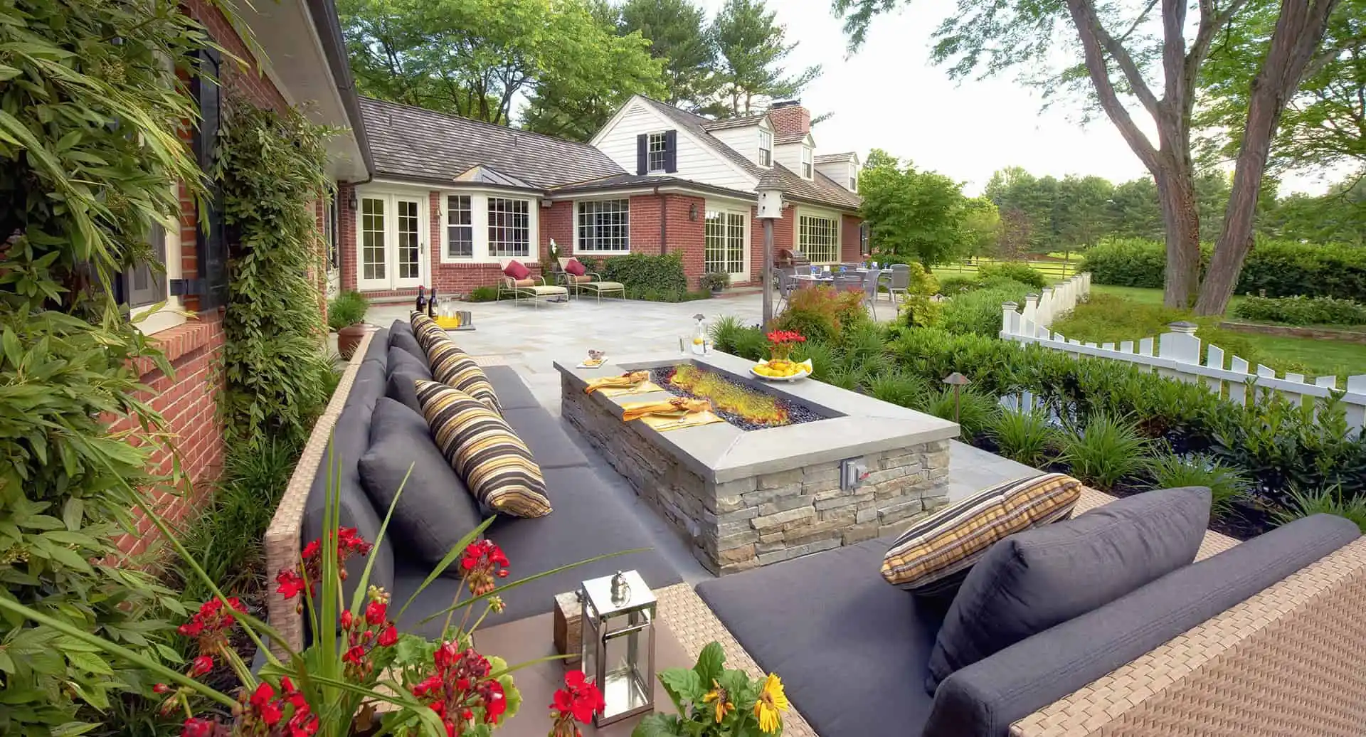 A patio with a stone fire pit surrounded by gray cushioned seating, striped pillows, and lush landscaping showcases exquisite outdoor living in Delaware County, PA. Red flowers adorn the foreground, while a brick house with large windows and a white fence completes the serene backdrop.