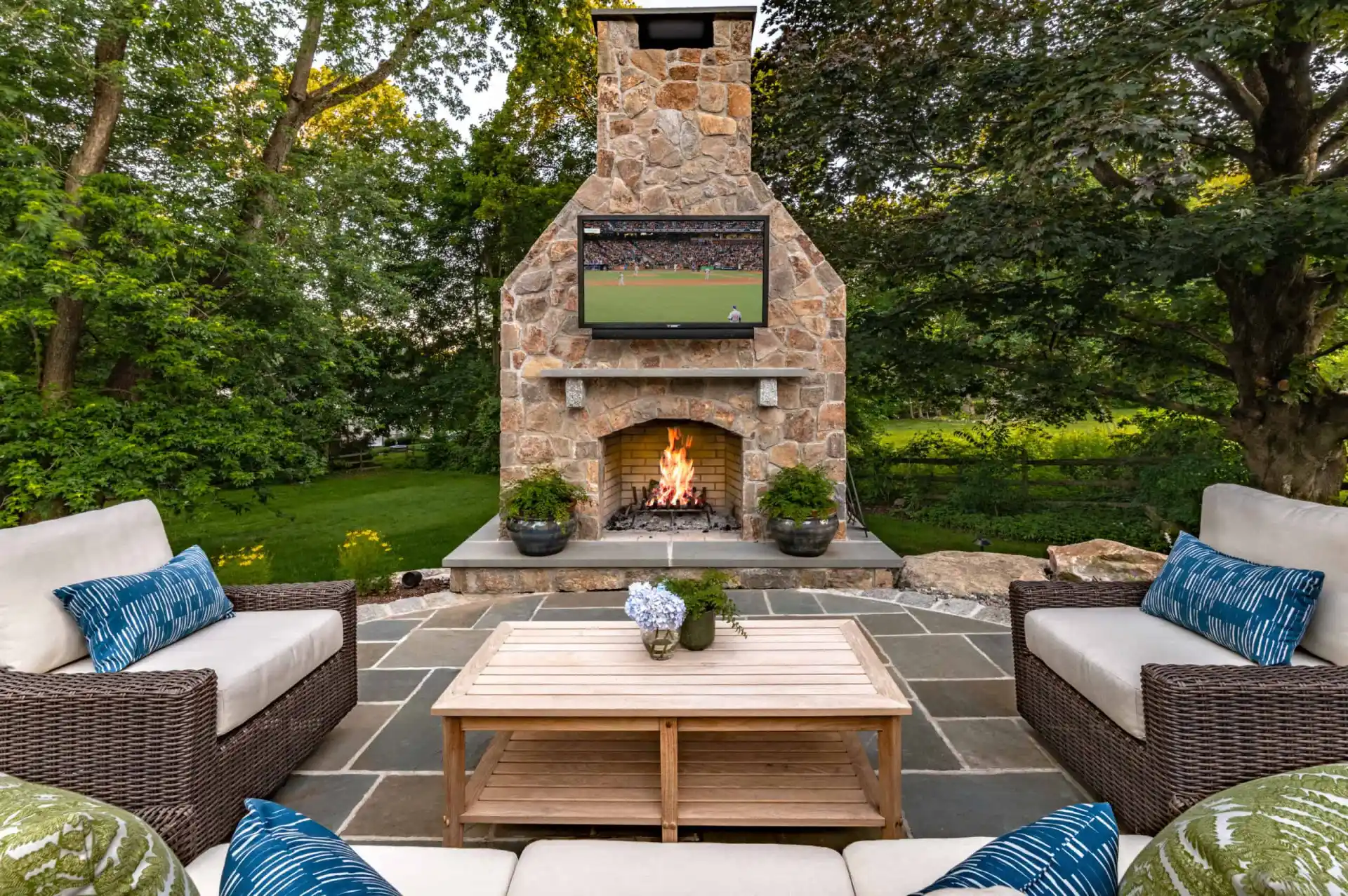 Enjoy outdoor living in Delaware County, PA, with a cozy patio featuring a stone fireplace and mounted TV displaying a thrilling sports game. Relax on cushioned wicker seating around a wooden coffee table adorned with flowers. The stone-tiled floor and lush green trees complete this luxurious retreat.