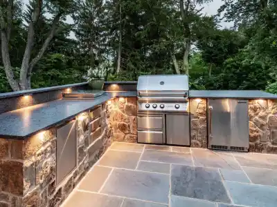 Outdoor living in Delaware County, PA, comes to life with a beautifully designed outdoor kitchen. Featuring stone countertops and cabinets with a stainless steel grill, mini fridge, and storage units on a stone patio. Built-in lighting enhances the space, surrounded by lush greenery.