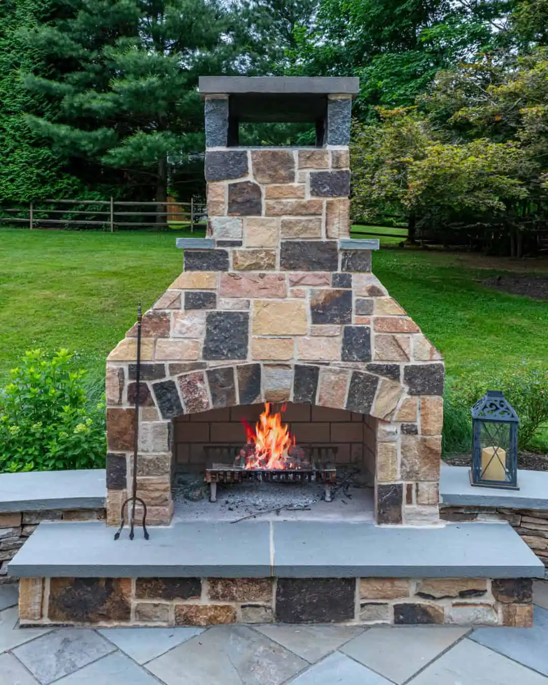 A cozy outdoor living scene in Delaware County, PA, features five Adirondack chairs around a fire pit, embraced by lush greenery and yellow flowers. String lights dangle overhead while houses and trees fade into the dusk backdrop.